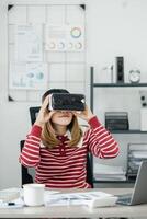 Woman with blonde hair in a striped sweater adjusts her virtual reality headset at an office desk with graphs on the wall. photo