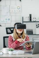 In an office setting, Woman donning a VR headset is simultaneously working with a tablet and a laptop. photo