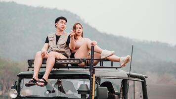 Young couple sits atop a safari vehicle, casually dressed, enjoying the view of a hazy, mountainous landscape. photo