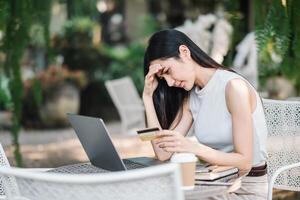 preocupado joven mujer experimentando problemas con un en línea pago, participación un crédito tarjeta y mirando a ordenador portátil pantalla a un al aire libre cafetería. foto