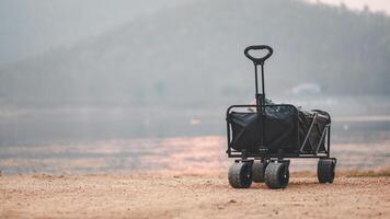 A black utility cart stands alone on a sandy lakeshore, against a backdrop of soft twilight hues. photo