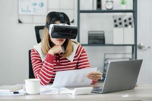 mujer en virtual realidad auriculares examina papel gráficos a un moderno trabajo escritorio con ordenador portátil y café. foto