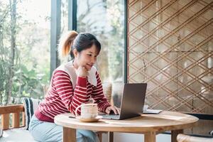 Cheerful woman enjoys a productive day working on her laptop in a well lit, plant-filled cafe, exuding a sense of satisfaction and comfort with a warm beverage by her side. photo