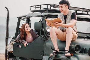 young woman laughs joyfully beside a man reading from a notebook, both perched on the hood of a safari car. photo