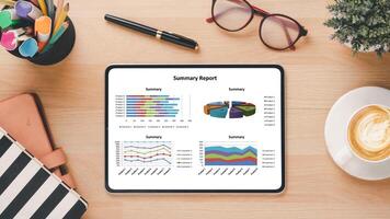 Overhead view of a tablet on a desk showing colorful summary reports, accompanied by office supplies, glasses, and a cup of coffee. photo