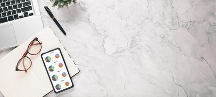 An organized marble desk featuring a modern laptop, a tablet with colorful pie charts, glasses, and a notebook for efficient business analysis. photo