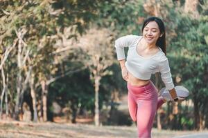 A smiling young woman performs a leg stretch, balancing on one foot in a sunny park, highlighting the importance of flexibility in fitness routines. photo