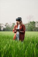 Woman holds a tablet and wears a virtual reality headset, standing in a lush green field with distant trees. photo