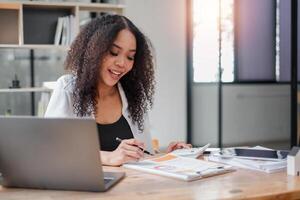 un optimista mujer de negocios con Rizado pelo sonrisas mientras trabajando en financiero cálculos, utilizando un calculadora y ordenador portátil a su de madera oficina escritorio. foto