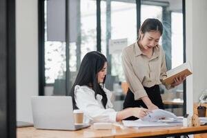 Two professional women are engaged in collaborative work, one reviewing documents while the other consults a notebook, in a well-lit modern office. photo