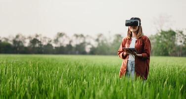 Woman holds a tablet and wears a virtual reality headset, standing in a lush green field with distant trees. photo