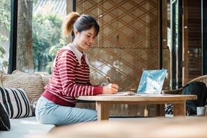 Happy young woman taking notes from her laptop in a cozy cafe corner surrounded by pillows and natural light. photo