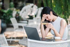 preocupado joven mujer experimentando problemas con un en línea pago, participación un crédito tarjeta y mirando a ordenador portátil pantalla a un al aire libre cafetería. foto