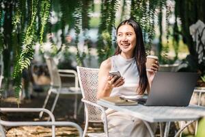 A joyful woman multitasks with a smartphone and coffee in hand, her laptop open on the table, all set against the lush backdrop of an outdoor garden workspace. photo