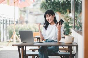 Joyful young woman celebrating success with smartphone and laptop at an open-air cafe, embodying achievement and happiness. photo