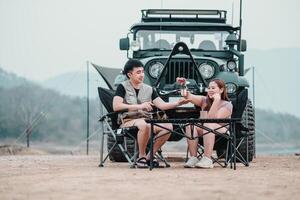 Man and woman share a cozy coffee break by a lake, with a rugged off-road vehicle and camping tent in the background. photo