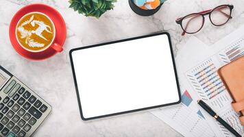 A professional workspace scene featuring a blank tablet screen, a cup of latte art coffee in a red mug, glasses, and financial charts, all arranged on a marble surface. photo