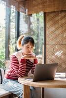 A focused woman sits absorbed in her work on a laptop at a round wooden table, surrounded by the comfort of a well-lit, plant filled room. photo