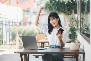 A young woman triumphantly raises her fist while looking at her smartphone, a gesture of victory, with a laptop on the table in an open-air cafe setting. photo