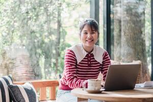 Cheerful woman looking camera and enjoys a productive day working on her laptop in a well lit, plant-filled cafe, exuding a sense of satisfaction and comfort with a warm beverage by her side. photo