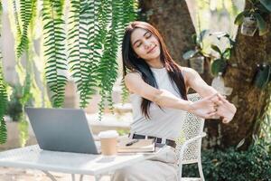 Relaxed female freelancer stretching arms while taking a break from work on her laptop in a lush garden cafe setting. photo