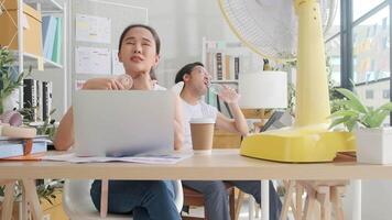 Two Asian workers are unhappy working in hot temperatures at desk in small business office in summer, uncomfortable with no air conditioner, blowing wind with fans, drinking water, and sweating. video