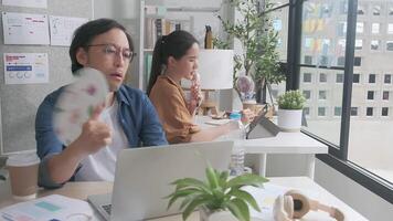 dos asiático trabajadores son infeliz trabajando con caliente temperaturas a casual oficina en verano estación, incómodo con No aire acondicionador, utilizando aficionados y Bebiendo agua, sentado a escritorios, y transpiración. video