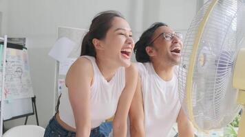 Two young Asian startup coworkers are fun blowing wind with electric fan, working in hot temperatures in a casual small business office in summer, refreshing from uncomfortable heat weather season. video