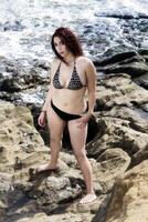 atractivo latina mujer en pie en bikini en rocas con Oceano foto