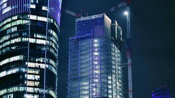 Skyscraper at construction process by night. Modern high-rise building under construction next to glass skyscrapers by night. photo