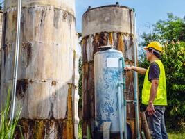 asiático hombre ingeniero controlador el calidad de agua lugares operando industrial agua purificación o filtración equipo antiguo cemento tanques para acuerdo agua en agua fábrica foto