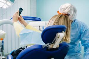 a dentist in a protective mask sits next to a patient and takes a selfie photo while working