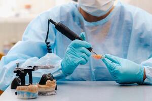 A masked and gloved dental technician works on a prosthetic tooth in his lab photo