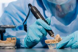 A masked and gloved dental technician works on a prosthetic tooth in his lab photo