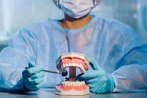 un dental médico vistiendo azul guantes y un máscara sostiene un dental modelo de el Superior y inferior mandíbulas y un dental espejo foto