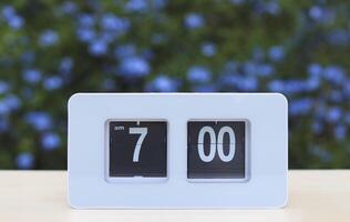 white flip clock  showing  7.00  o clock on the table in the garden with purple flowers background. photo