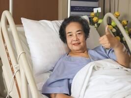 cheerful  Asian senior  female patient  lying in hospital bed,  smiling and showing her thumb up. Elderly health concept. photo