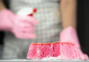 Kitchen sponge in soapy suds on the table. In the background in blur a woman in rubber gloves and with detergent. Close-up. Selective focus. photo