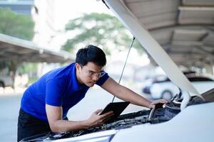 A man is looking at a tablet while working on a car. He is wearing a blue shirt and glasses photo
