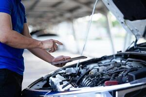 A man in a blue shirt is looking at a tablet while standing next to a car. He is checking the car's engine photo