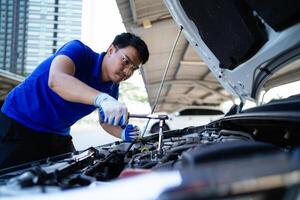 A man is working on a car engine. He is wearing a blue shirt and gloves. The car is parked under a building photo