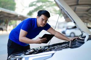 A man in a blue shirt is looking at a tablet while standing next to a car. He is checking the car's engine photo