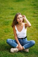 Beautiful young woman wearing tank top and jeans sitting on grass lawn photo