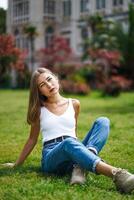 Beautiful young woman wearing tank top and jeans sitting on grass lawn photo