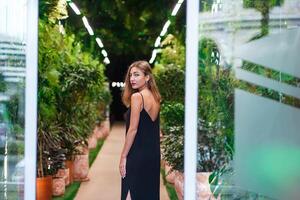 Young woman wearing black dress on thin straps stands among green plants inside arrangement photo