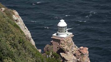 Visão do uma tranquilo branco farol em uma penhasco com exuberante vegetação, supervisionando a rítmico oceano ondas em uma ensolarado dia. video