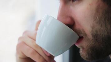 un' uomo bevande caffè di il finestra nel il mattina. avvicinamento di un' bianca tazza e labbra di un' uomo potabile caffè video