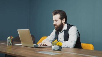 positif homme avec une barbe travail sur une portable et la programmation, Cordialement en riant à la recherche à le caméra. positif employé en riant à travail dans le bureau. video