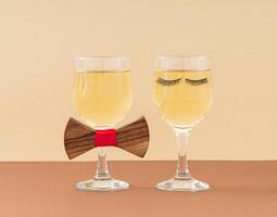 Romantic layout with two glasses of white wine, eyelashes and bow tie on brown and beige background. People lifestyles and relationships in love concept. Fashion aesthetic party or celebration idea. photo