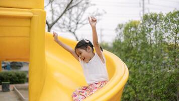 A young girl is sitting on a yellow slide, smiling and waving photo